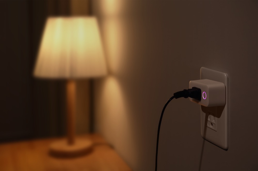 間接照明のスタンドをつないだ SwitchBot プラグミニ。壁のコンセントに挿さり、正面のボタンが紫色に点灯している